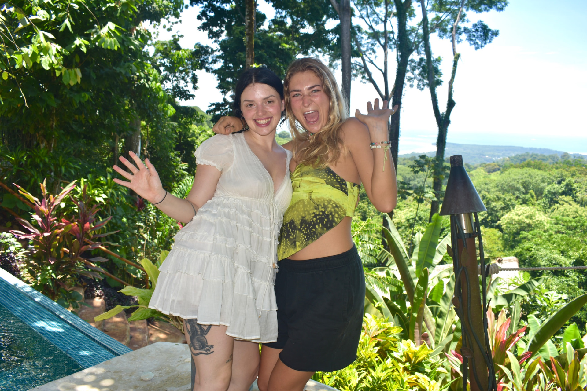 Two friends waving beside the infinity pool with the ocean view behind